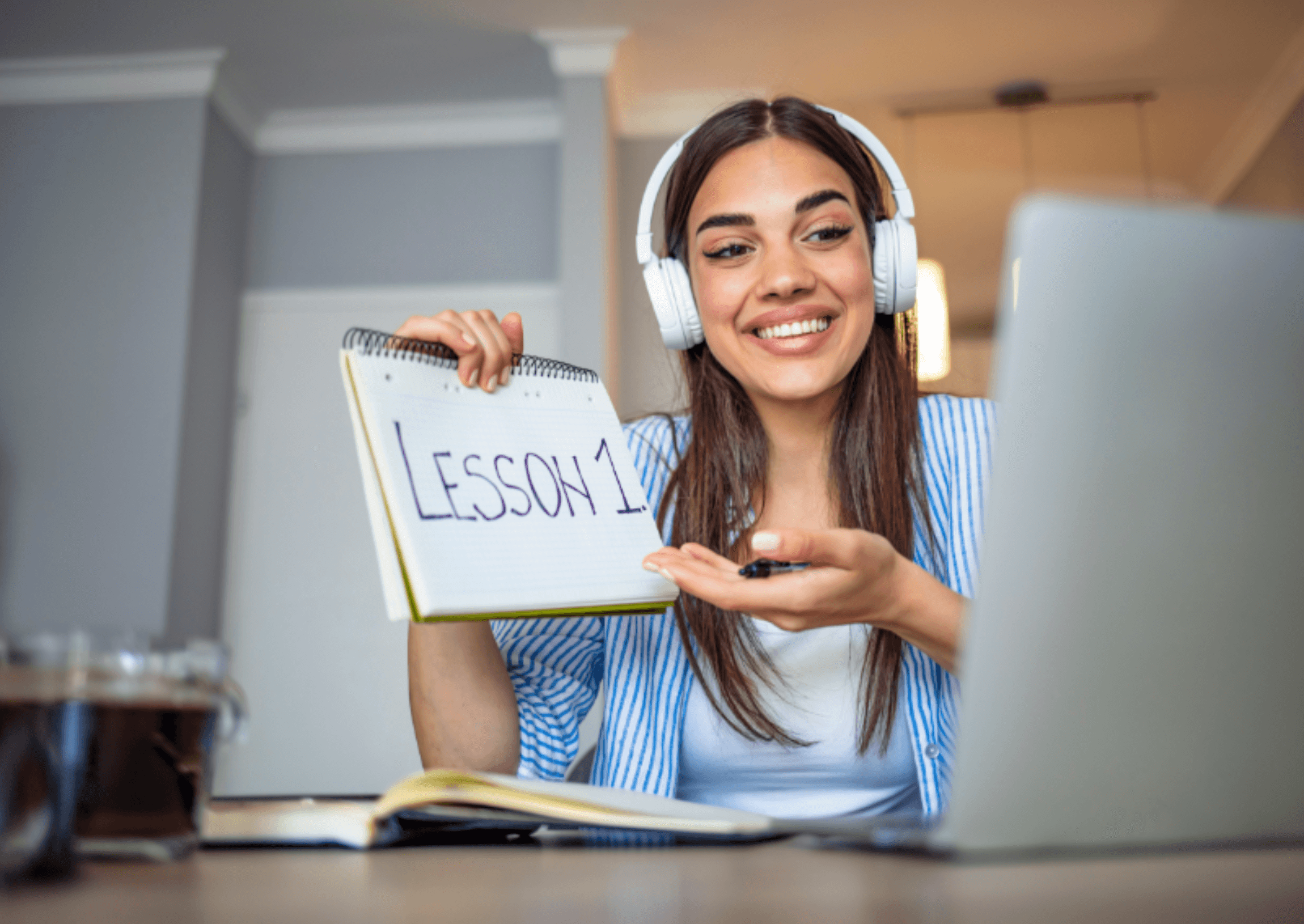 A tutor in headphones holding up a notepad that reads Lesson 1 to her laptop